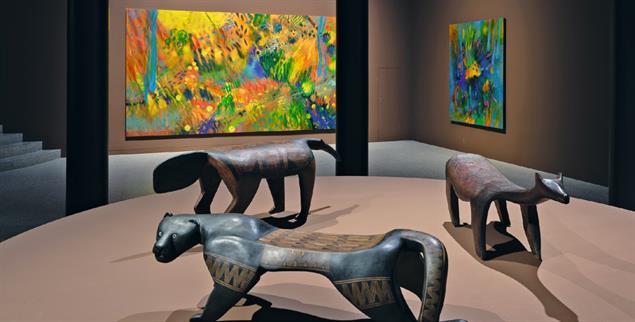 Bunte Welt: Vor den Gemälden von Carlos Jacanamijoy stehen drei moderne Tierbänke aus dem Xingu-Gebiet im Kreis (Foto: Simon Vogel, 2026 © Kunst- und Ausstellungshalle der Bundesrepublik Deutschland)